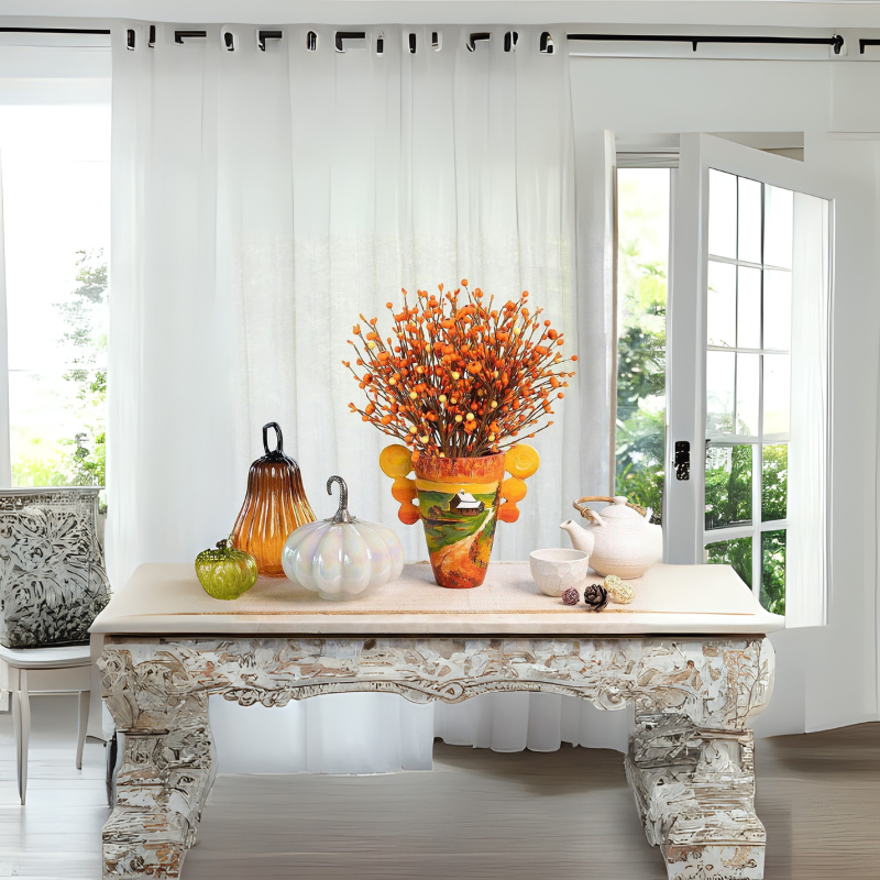 Elegant autumn-inspired table décor from Grella Boutique featuring a vibrant orange floral arrangement in a decorative vase, glass pumpkin ornaments in amber, pearl, and green, alongside a white teapot set on an ornate carved wooden table.