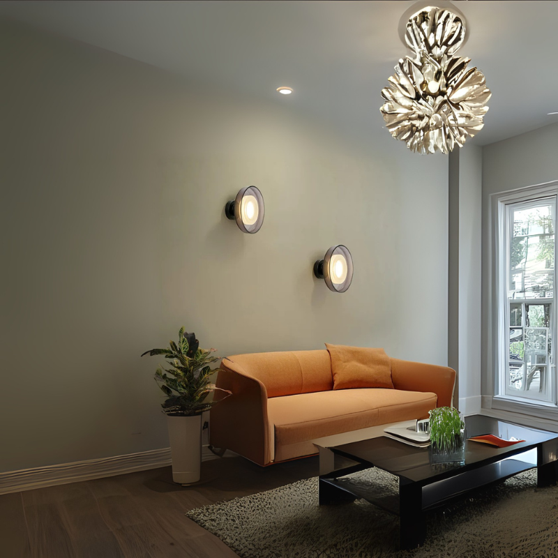Contemporary living room from Grella Boutique featuring a sleek orange sofa, modern black coffee table with décor accents, potted indoor plant, wall-mounted circular lights, and a statement ceiling light fixture near a large window.